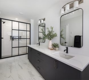 Primary ensuite with a large shower with sliding glass doors trimmed with black, two square sinks with a beige quartz counter, black single handle faucets, dark wood vanity, two rounded and black trimmed mirrors. The flooring is marble-like 12x24 tiles.