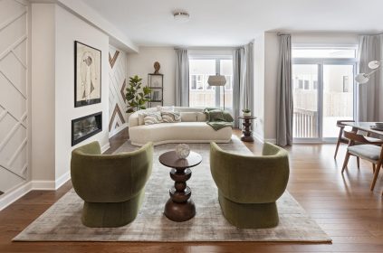 A modern living room featuring two green chairs facing a beige sofa on a large rug. The space is decorated with abstract art, plants, and a small side table. Large windows allow natural light in, illuminating the wooden floor and neutral-toned decor.and large window in the living space complete the bright, airy look.