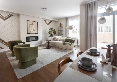 A modern living room featuring two green chairs facing a beige sofa on a large rug. The space is decorated with abstract art, plants, and a small side table. Large windows allow natural light in, illuminating the wooden floor and neutral-toned decor.