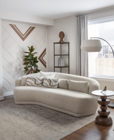 Modern living room with beige rounded sofa, a geometric accent wall featuring wood and white details, a potted plant and wood and black metal shelf unit, a beige floor lamp and a small bird on a side table.