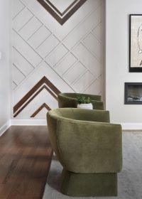 Modern living room with two olive-green velvet chairs, a geometric accent wall featuring wood and white details, a small plant on a side table, and part of an electric fireplace