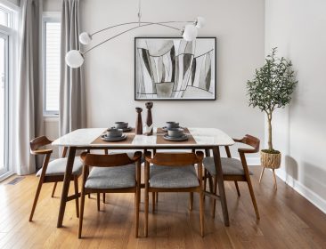 A modern dining room with a wooden table and six chairs. The table is set with decorative items. A contemporary light fixture hangs above. Theres a large abstract painting on the wall and a potted plant in the corner. Hardwood flooring complements the space.