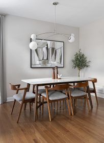 A modern dining room with a wooden table and six chairs. The table is set with decorative items. A contemporary light fixture hangs above. Theres a large abstract painting on the wall and a potted plant in the corner. Hardwood flooring complements the space.