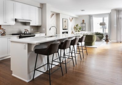 Modern open-concept Kitchen and Great Room living area with a white island, brown leather bar stools, stainless steel appliances, and hardwood floors. A green armchair and cream sofa and large window in the living space complete the bright, airy look.