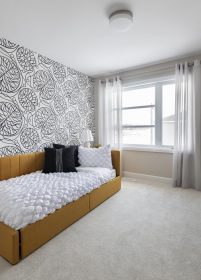 A bedroom features a single bed with a mustard-yellow frame and white bedding. The wall behind the bed has a black and white abstract patterned wallpaper. A large window with sheer curtains allows natural light to fill the room.