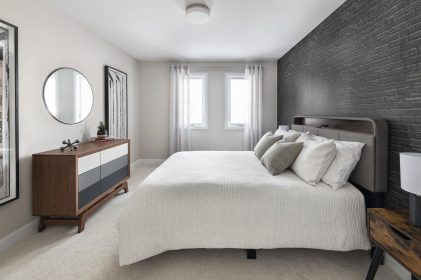 Secondary bedroom with large bed with beige textured beige bedding. The wall behind the bed features faux dark grey bricked wallpaper. There is a round mirror over the wood dresser with multi grey drawers flanked by tall, framed hockey stick artwork on each side. There are similar wood nightstands on each side of the bed with black lamps and white shades. The room features beige carpeting. Sheer curtains allow lots of natural light from the large window in.