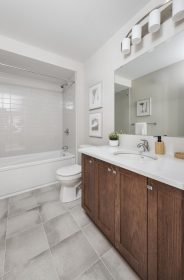 Modern bathroom with a brown oak cabinet vanity, white quartz countertop, and large mirror. Features a toilet beside a shower-tub with neutral tiles cover the floor and walls, complemented by framed artworks and soft lighting.