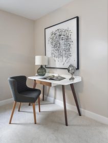 White desk with wooden legs and gray glass and metal accessories and an open book. There is framed artwork on the wall and an upholstered gray chair with brown wooden legs.