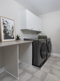 Laundry room with beige 12x12 tile flooring, laminate countertop, a steel-coloured front load washer and dryer with towels on top and a set of white melamine upper cabinets with 4 doors.