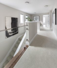 Second floor beige carpeted upper hall area featuring art on the walls and a clear glass wall opening with chrome hardware where the staircase is visible behind it. There are two windows in the stairwell .