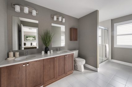 Modern bathroom with dual sinks on an oak vanity, large dual mirrors, and vanity lighting. A plant decorates the white quartz countertop. A toilet is beside the vanity, and a glass-enclosed shower is on the right. Natural light streams through a window with blinds.