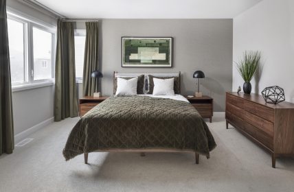 A modern bedroom with a green textured bedspread, two matching wooden nightstands with black lamps, and a dresser on beige carpeting. Large windows with green curtains provide natural light, and a geometric artwork hangs above the bed. A plant decorates the dresser.