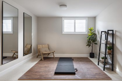 A minimalist home gym in the basement with a yoga mat with a faux wood foam floor tiles on beige carpet. Two large mirrors are mounted on the left wall. A wicker chair and a potted plant are in the corners, and a black shelving unit with folded towels stands to the right. There is a large basement window that lets in lots of natural light.