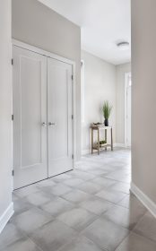 A modern front foyer/entryway with light gray tiled flooring, beige walls, and a double door closet with lever handles on the left. A wooden console with decorative items and a tall plant is near the glass front door at the front of the hallway.