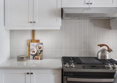 A modern kitchen with quartz counter featuring a cookbook and a cutting board, with a gas stove and a metallic kettle on the right. White cabinetry and light-colored tiled backsplash add to the clean and minimalist design.